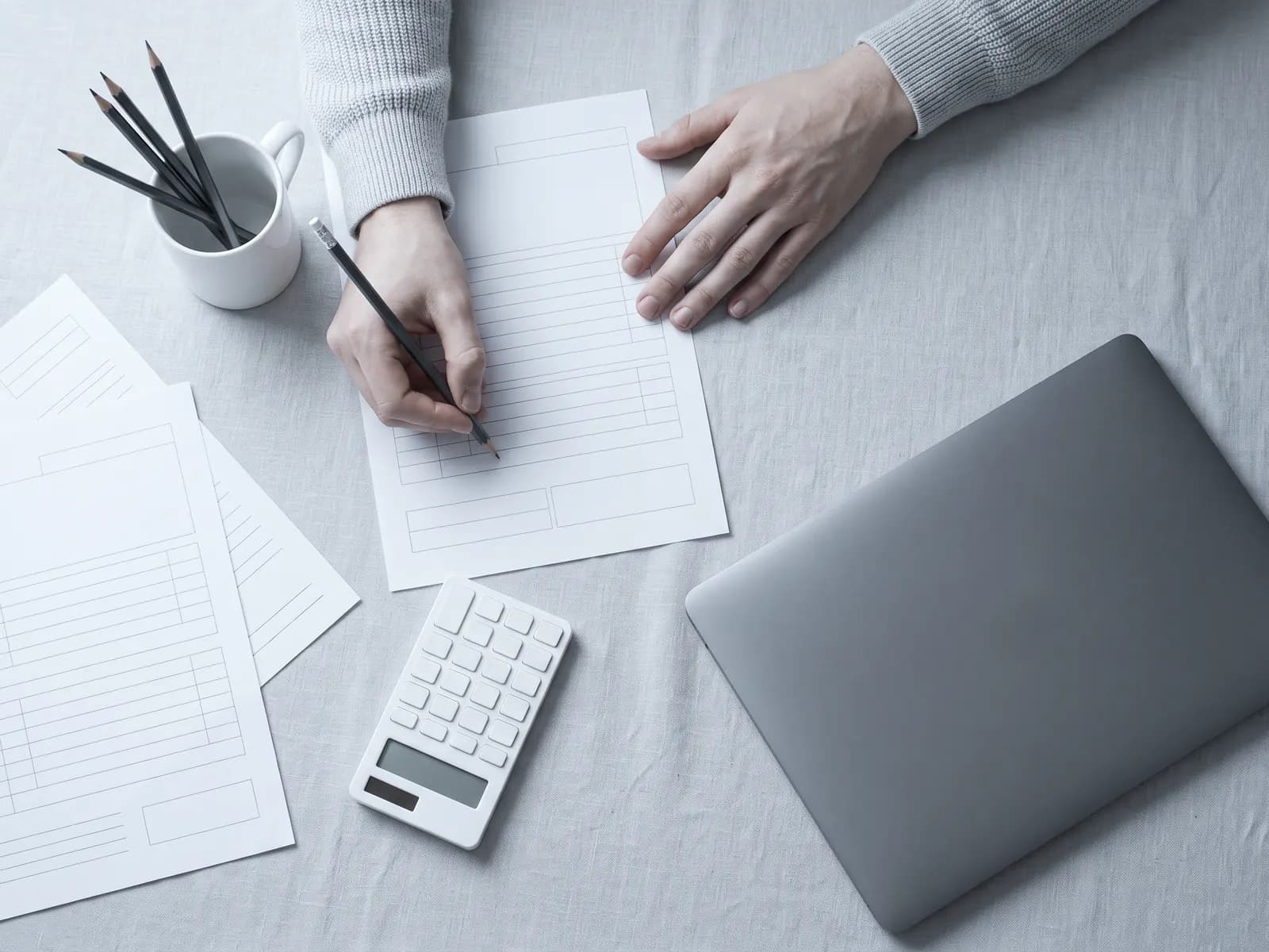 Hands at a bookkeeper’s desk: a pencil over printed form pages, a calculator and a closed laptop, in cool daylight.